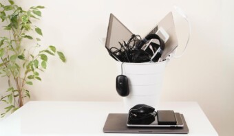 Un cubo blanco repleto de dispositivos electrónicos y cables, entre ellos un ordenador portátil y unos auriculares, simboliza discretamente la obsolescencia mientras reposa sobre una mesa. Un ratón de ordenador cuelga del cubo, mientras que plantas y otros aparatos electrónicos permanecen en el fondo.