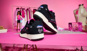 Las zapatillas Reebok con temática de Barbie se exhiben sobre una mesa rosa rodeada de artículos de moda y un maniquí.