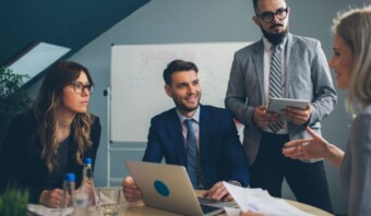 Cuatro profesionales se encuentran enfrascados en una discusión en un entorno de oficina. Con una pizarra de fondo, se reúnen alrededor de una mesa equipada con un portátil y documentos, centrándose en el blindaje corporativo estratégico para fortalecer su enfoque empresarial.