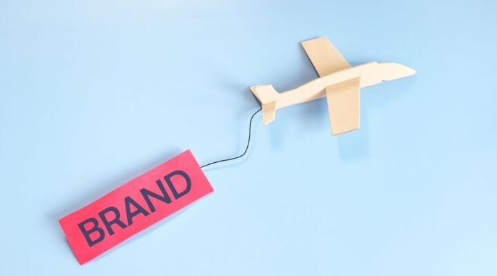 Un avión de papel se desliza sobre un fondo azul claro, remolcando una etiqueta roja que dice "MARCA", que recuerda a un tablero de marca en movimiento.