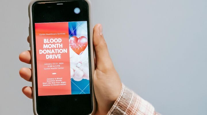 Una persona sostiene un teléfono que muestra un cartel de la campaña de donación del Mes de Sangre con detalles vibrantes del evento, que incluyen fecha, hora y ubicación sobre un fondo rojo llamativo: una campaña publicitaria impactante.