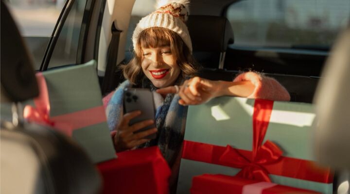 Una mujer en un automóvil, sonriendo a su teléfono, está rodeada de regalos envueltos, abrazando el espíritu de fin de año.