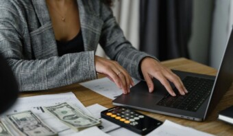 Una persona trabajando en un escritorio usando una computadora portátil, con papeles, una calculadora y billetes de dólares estadounidenses esparcidos por todos lados.