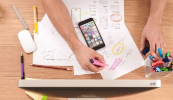 Una persona escribe con resaltadores en papeles llenos de bocetos y notas, desarrollando una idea, mientras un teléfono inteligente muestra una cuadrícula de aplicaciones cerca. Varios bolígrafos y marcadores están esparcidos sobre un escritorio de madera junto a un monitor de computadora.