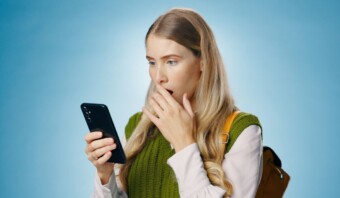 Una mujer con cabello largo parece sorprendida, posiblemente después de encontrarse con publicidad engañosa, mientras sostiene un teléfono inteligente sobre un fondo azul.