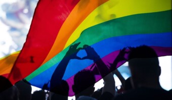 Varias siluetas de personas sostienen una bandera de arcoíris sobre sus cabezas, con sus manos formando un corazón debajo de ella.