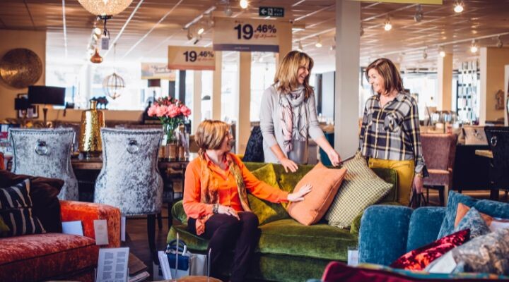 Tres mujeres entablan una animada conversación mientras eligen cojines para sofás de colores en una tienda de muebles. La iluminación brillante y los diversos elementos de decoración del hogar en el fondo crean una atmósfera acogedora que recuerda a las atractivas estrategias de conversación de marketing. marketing conversacional