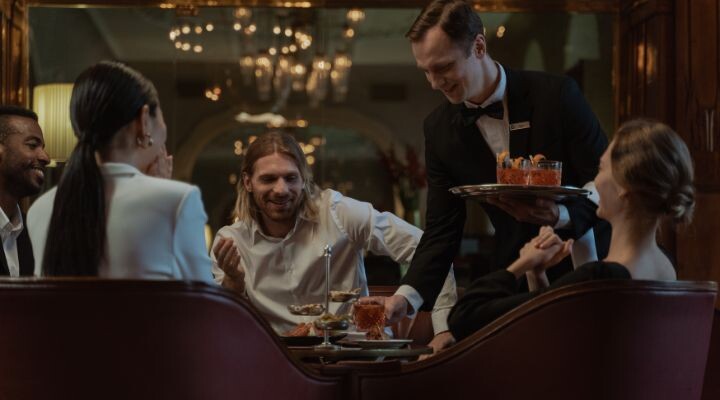 Un camarero sirve comida a cuatro personas en una mesa de restaurante, rodeado de una elegante decoración y una lámpara de araña, realzando la experiencia general con elementos de marketing gastronómico que tientan tanto el gusto como la atmósfera. marketing auditivo