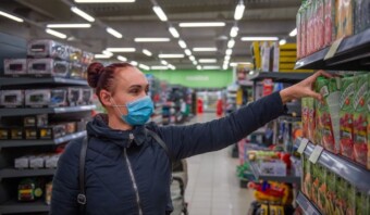 En un supermercado lleno de gente, una mujer con una máscara y una chaqueta azules alcanza un artículo en el estante, con pasillos de juguetes coloridos que recuerdan a los que se encuentran en México y Brasil en el fondo.