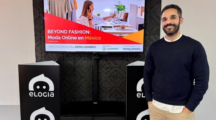 Una persona se encuentra junto a una pantalla que muestra una presentación titulada "Más allá de la moda: Moda Online en México, octubre de 2023", con la marca eLogia en los podios.