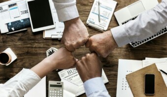Cuatro manos de diversos tonos de piel chocan los puños sobre una mesa con papeles, una calculadora, una tableta y una taza, lo que significa trabajo en equipo o colaboración.