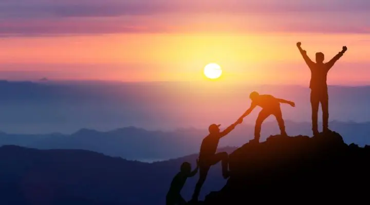 Siluetas de tres personas escalando una montaña al atardecer; uno ayuda a otro a subir mientras el tercero celebra en la cima.