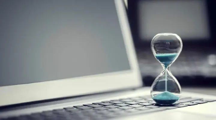 Un reloj de arena con arena azul reposa sobre el teclado de una computadora portátil, con una pantalla borrosa en el fondo.