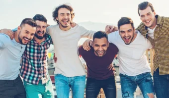 Seis hombres vestidos con ropa informal se encuentran muy juntos, sonriendo y apoyándose uno en el otro frente a un fondo al aire libre, celebrando el Día del Hombre con camaradería y alegría.