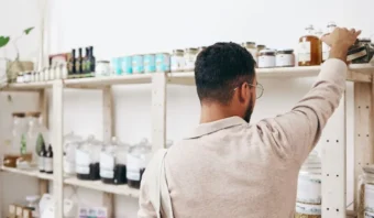 En 2025, una persona con gafas y un suéter beige alcanza un producto en un estante de una tienda bien abastecido, donde varios frascos y botellas brillan de manera tentadora.