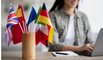 Una persona está sentada en un escritorio con un ordenador portátil, concentrada en buscar oportunidades laborales. A su lado, un jarrón de madera sostiene varias banderas internacionales pequeñas, incluidas las del Reino Unido, España, Francia y Alemania.