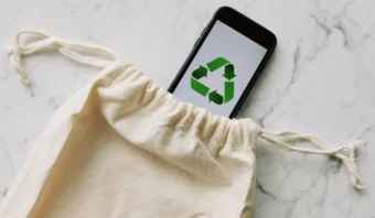 Un teléfono inteligente que muestra un símbolo de reciclaje en su pantalla se encuentra parcialmente colocado dentro de una bolsa de tela con cordón, descansando elegantemente sobre una superficie de mármol. Esta escena subraya sutilmente las preferencias verdes, combinando tecnología moderna con valores ecológicos.