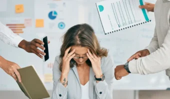 Una mujer se sienta en un escritorio con la cabeza entre las manos, encarnando los desafíos de la "salud mental en la búsqueda de empleo", rodeada de personas que sostienen documentos, un teléfono y una tableta, todo ello con un telón de fondo de gráficos y notas adhesivas.