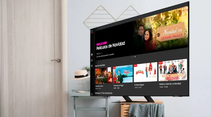Un televisor Samsung TV Plus muestra una selección de películas y series navideñas tituladas "Películas de Navidad". Cerca de allí, una mesa adornada con un sombrero y una decoración festiva completa la acogedora escena.