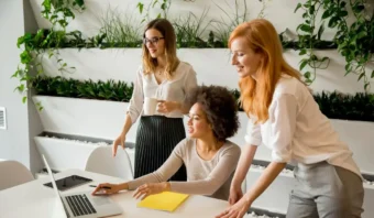 Tres empresarias mexicanas colaboran en una oficina, rodeadas de una computadora portátil, un cuaderno y plantas en la pared.