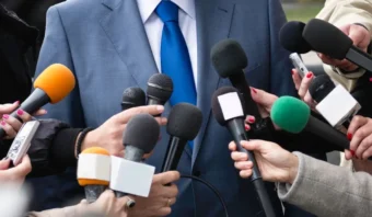 Una persona con traje y corbata azul, que irradia la confianza que da la formación de un periodista, está rodeada de múltiples micrófonos sostenidos por varias personas, lo que sugiere una conferencia de prensa o una entrevista con los medios.