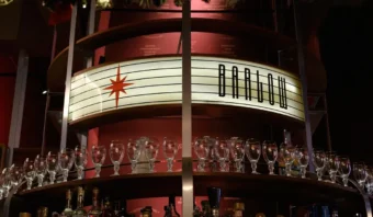 Un estante de bar con hileras de vasos y botellas vacíos. Un cartel en el estante dice "BARROW en CDMX" con un diseño de estrella roja.