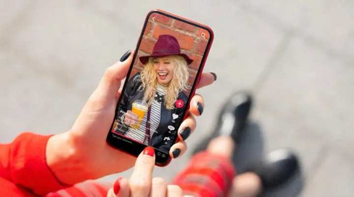 Una persona sostiene un teléfono inteligente que muestra a un individuo sonriente con un sombrero, bebiendo casualmente una bebida frente a una pared de ladrillos rústicos de fondo, cautivando a audiencias generacionales con su atractivo atemporal.