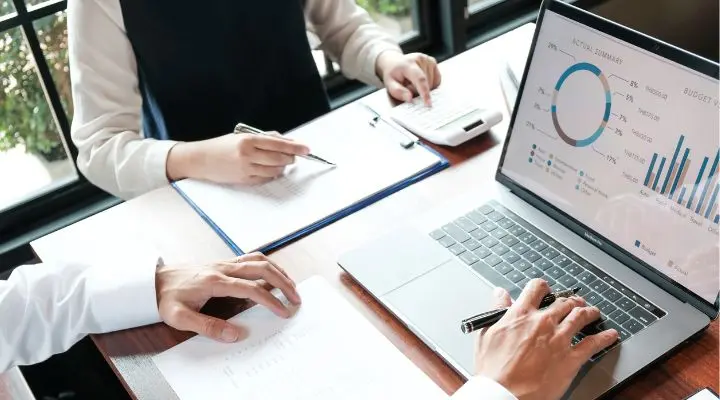 Dos personas trabajan en una mesa, una con un ordenador portátil que muestra gráficos relacionados con el análisis de mercado y la otra escribiendo en un bloc de notas. También se ve una calculadora.