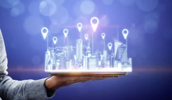 Una mano sostiene una tableta digital que muestra un paisaje urbano futurista mejorado mediante geolocalización, con marcadores de ubicación flotantes sobre un fondo azul.