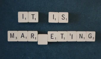 Las fichas de Scrabble escriben "IT IS", con "MARKETING DE PRODUCTO" debajo sin la letra "K" sobre un fondo azul oscuro.
