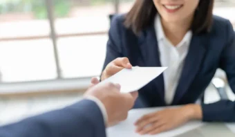 Una persona con traje le entrega un sobre a otra persona sentada, que está sonriendo.