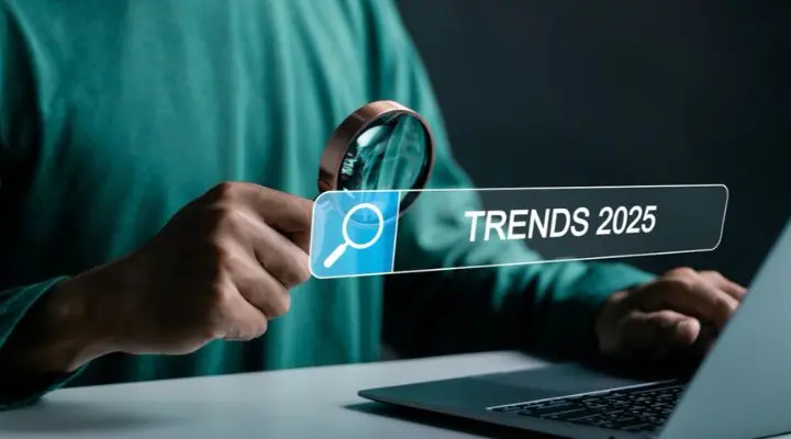 Una persona sostiene una lupa sobre una barra de búsqueda digital que muestra "Tendencias 2025" junto a una computadora portátil, capturando perfectamente la esencia de la mezcla de medios en el cambiante panorama digital.