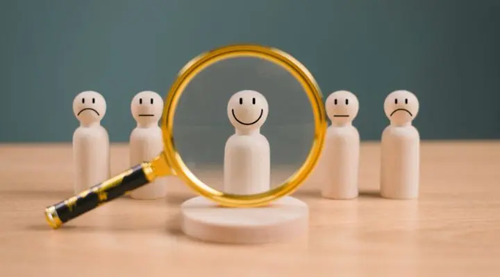 Figuras de madera con distintas expresiones faciales se encuentran sobre una mesa, creando una dinámica mezcla de medios. Una lupa enfoca una figura con un rostro sonriente, resaltando la expresiva artesanía.
