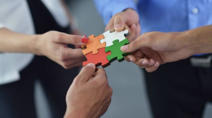 Cuatro manos que sostienen piezas de rompecabezas conectadas en naranja, tostado, verde y blanco, que simbolizan el trabajo en equipo y la colaboración como parte de una estrategia de comunicación.