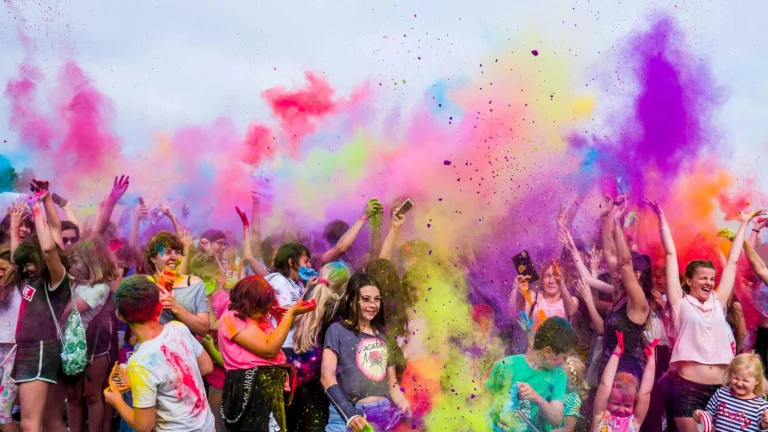 Una multitud de personas celebra en un festival, lanzando polvos de colores vibrantes al aire, creando una atmósfera animada con nubes de tonos púrpura, amarillo y rosa: un ejemplo perfecto de marketing experiencial.