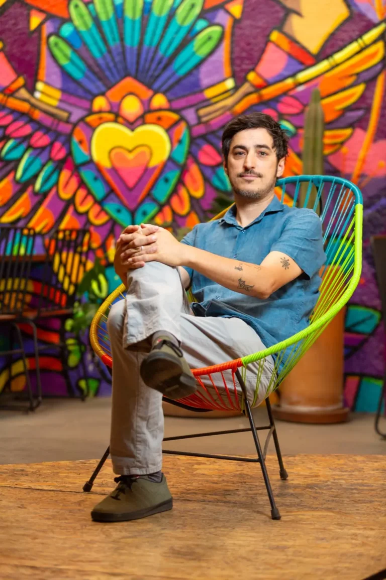 Una persona se sienta con las piernas cruzadas en una colorida silla Campo Vivo sobre un fondo mural vibrante que presenta diseños abstractos de corazones y plumas.