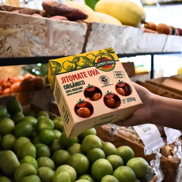 Una mano sostiene una caja de tomates uva orgánicos Campo Vivo etiquetados como "Jitomate Uva", colocados encima de una pila de productos verdes en una concurrida tienda de comestibles.