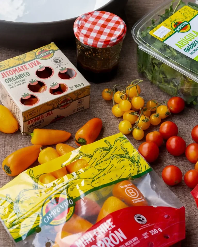 Sobre la mesa se encuentran elegantemente surtidos tomates Campo Vivo en distintos envases, un tarro con tapa roja y blanca y un recipiente con rúcula.