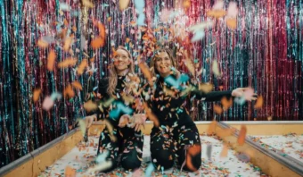Dos personas se arrodillan y lanzan confeti de colores al aire. Están rodeadas de serpentinas brillantes en el fondo.