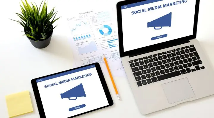 Dos dispositivos muestran la palabra "Social Media Marketing" con un icono de megáfono, destacando las campañas integrales en redes sociales. Cerca de allí, una planta, documentos con gráficos, un lápiz y una nota adhesiva descansan sobre la superficie blanca.