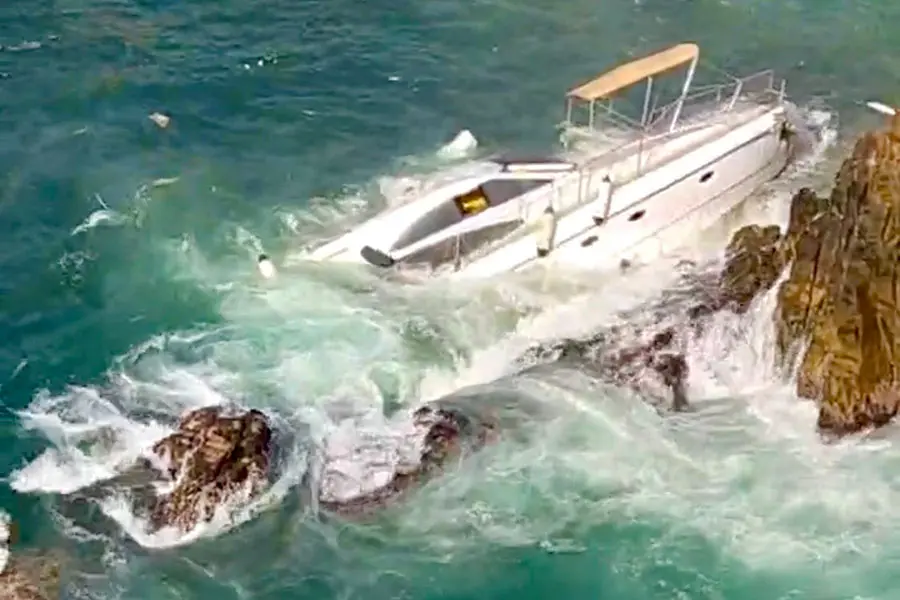 Un yate blanco está varado en rocas irregulares en aguas turbulentas y espumosas bajo un cielo despejado.
