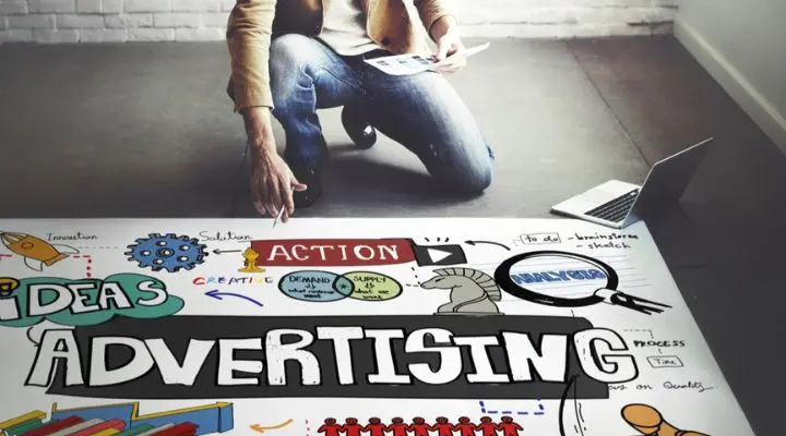 Una persona se arrodilla junto a una mesa adornada con un cartel publicitario de colores. Sobre la mesa hay un ordenador portátil y papeles que enfatizan conceptos como "ideas", "acción" y "solución".