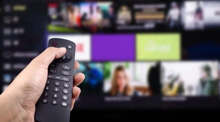 Una mano agarra un control remoto de televisión, colocada frente a una pantalla borrosa que muestra varios canales en una televisión conectada.