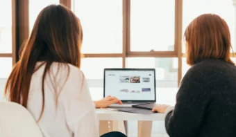 Dos personas sentadas en una mesa colaboran mientras observan la pantalla de un portátil que muestra diversas imágenes y contenidos. La escena se desarrolla en una habitación bien iluminada con grandes ventanales al fondo.
