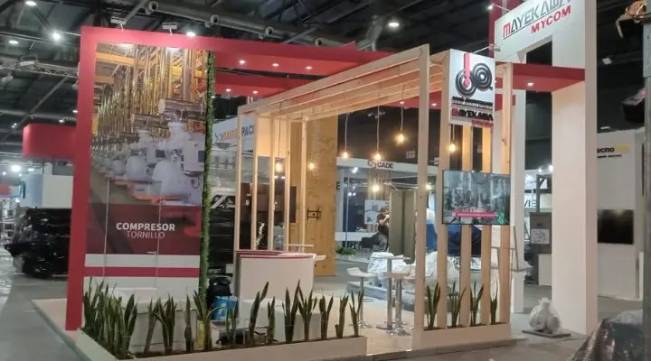 Stand de feria con estructura de madera, plantas e imágenes de maquinaria industrial. El cartel dice "Compresor Tornillo". Las mesas y sillas invitan a entrar en lo que realmente es el mejor espacio en una exposición.