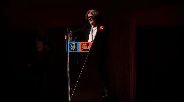 Una persona con el pelo largo habla de pie ante un podio transparente. El podio tiene dos logotipos de colores pegados. La iluminación es tenue y el fondo es oscuro celebrando el festival Circulo de oro 2025