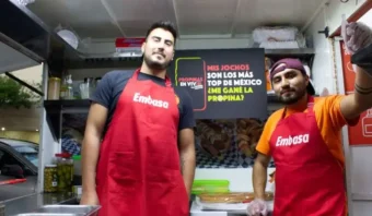 Dos hombres con delantales rojos de Embasa están de pie en un camión de comida. Un cartel detrás de ellos menciona la excelente calidad de los hot dogs mexicanos y las propinas.