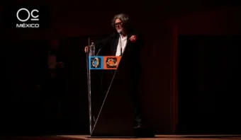 Una persona está de pie en un podio sobre un escenario oscuro y habla a través de un micrófono. El podio tiene paneles coloridos con logotipos.