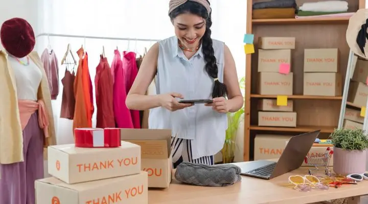 Una persona toma una foto de una prenda de vestir en una oficina en casa, disfrutando de la experiencia de vender en línea. Cajas de envío, ropa empaquetada, una computadora portátil y un estante de prendas vibrantes los rodean mientras perfeccionan la toma.