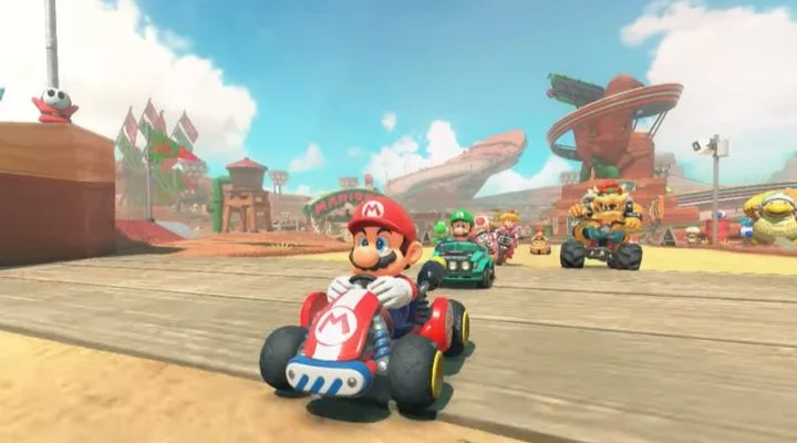 Personajes que compiten en karts en una pista de arena en un videojuego, con un paisaje vibrante y espectadores animando en el fondo.